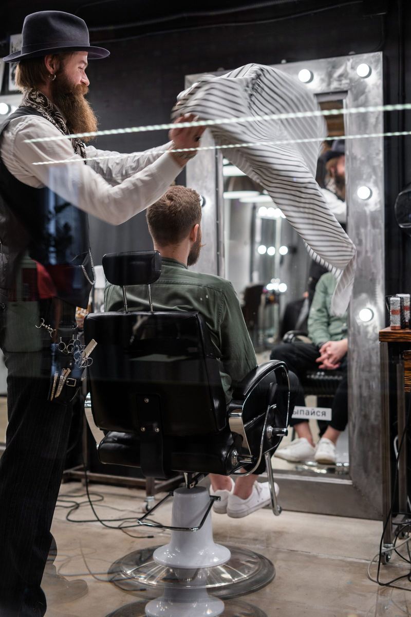A barber in a felt hat swings a striped cape over a client in the chair, mirror lights behind.