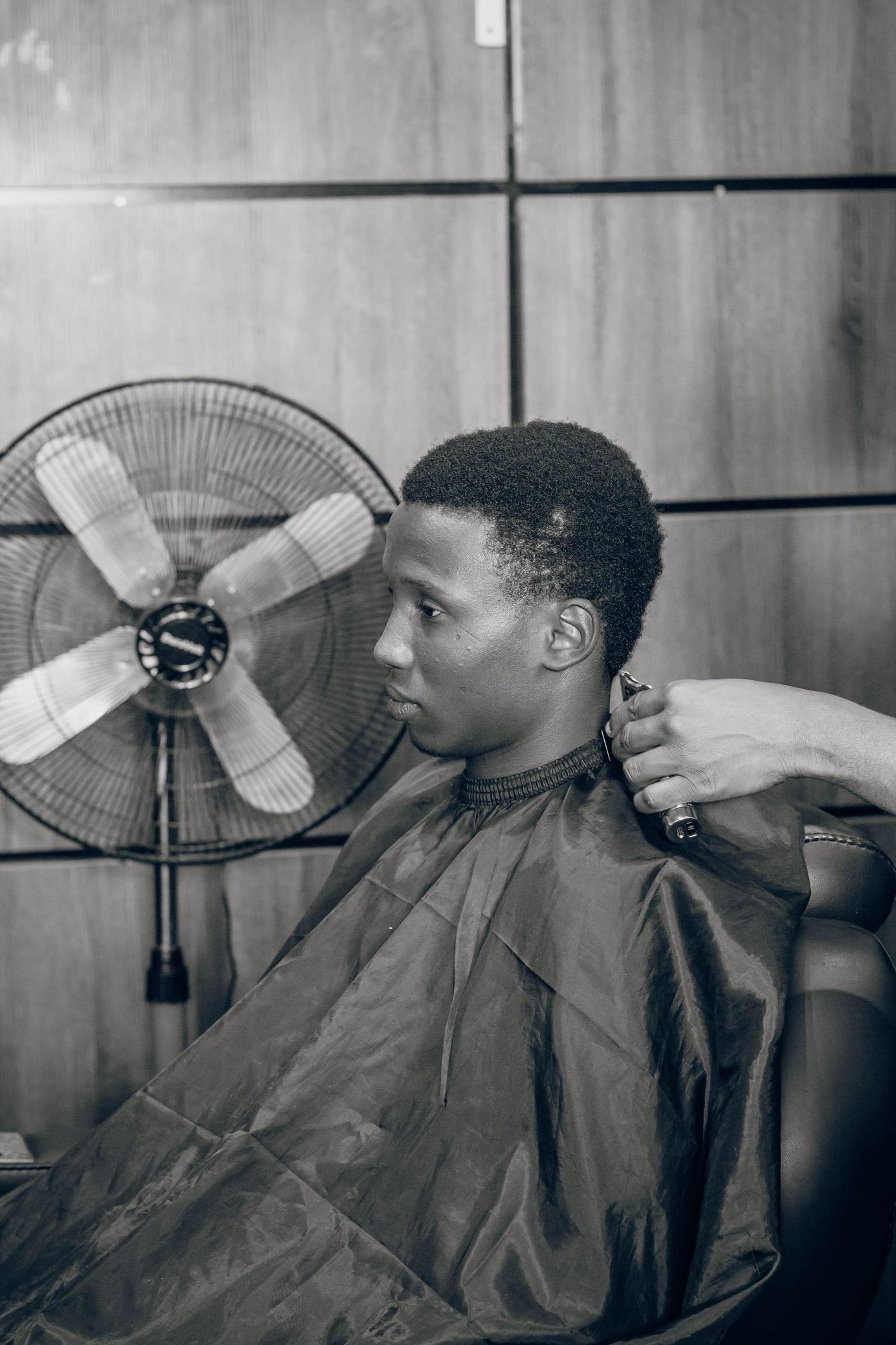 Black-and-white photograph: a barber's hand runs clippers along a caped client's neckline, a standing fan behind them.