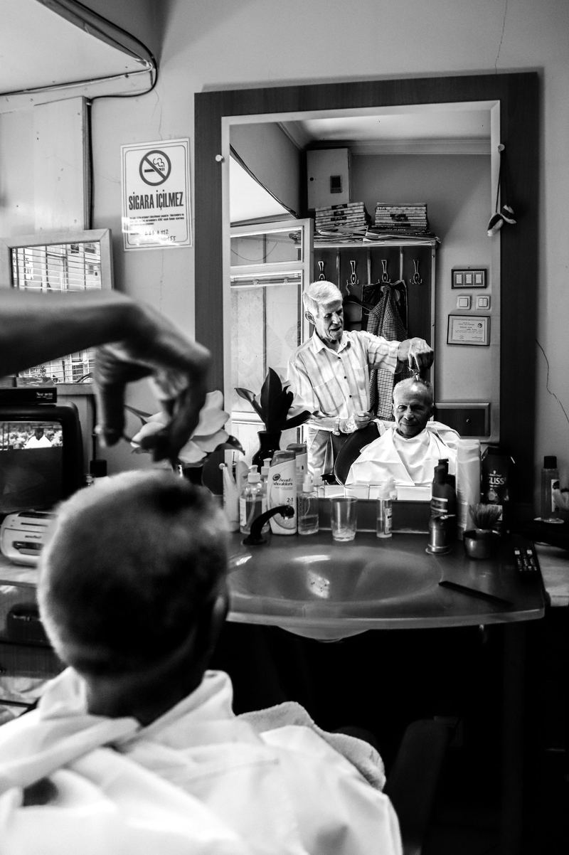 Seen past a client's shoulder, an older barber works on a seated customer, reflected in the mirror.