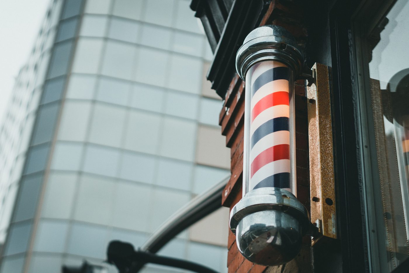 A lit red, white and blue barber's pole on an old shopfront, with a glass office tower rising behind it.