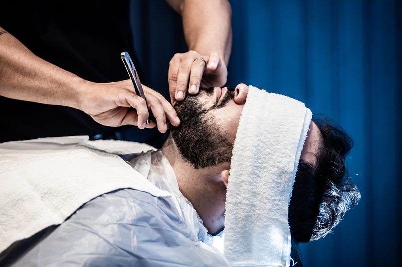 A barber shapes a client's beard with a straight razor, a white towel folded over his eyes.