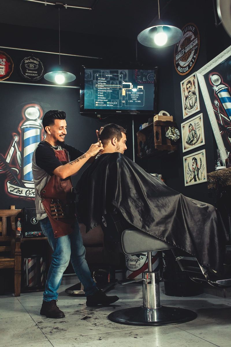 A barber in a dark apron cuts a caped client's hair in a shop hung with framed prints.
